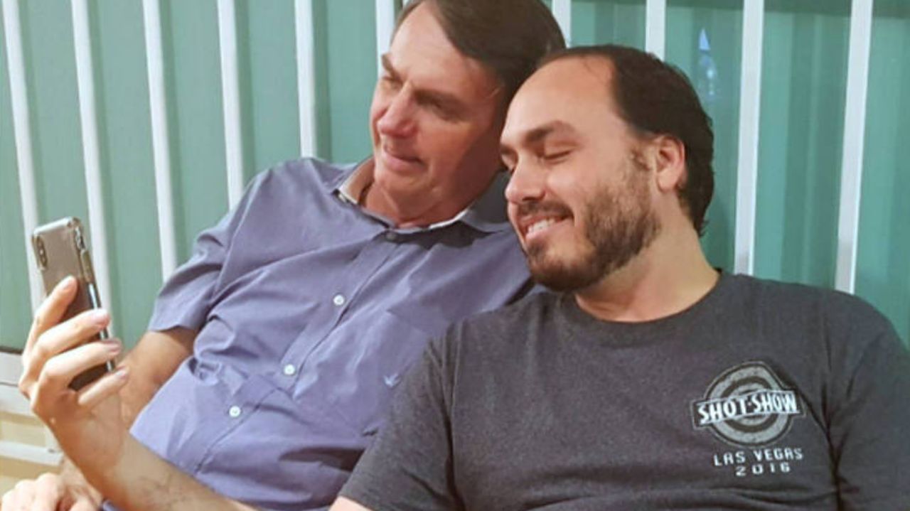 Carlos Bolsonaro e Jair Bolsonaro sorrindo e olhando para celular