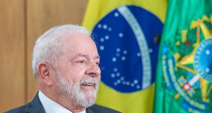 Lula sorrindo, de perfil, com bandeira brasileira de fundo