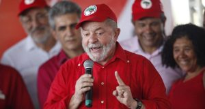 Lula com camisa vermelha e boné do MST, falando em microfone e gesticulando