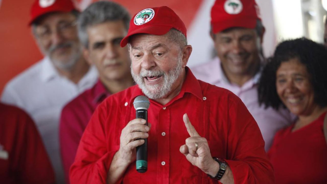 Lula com camisa vermelha e boné do MST, falando em microfone e gesticulando