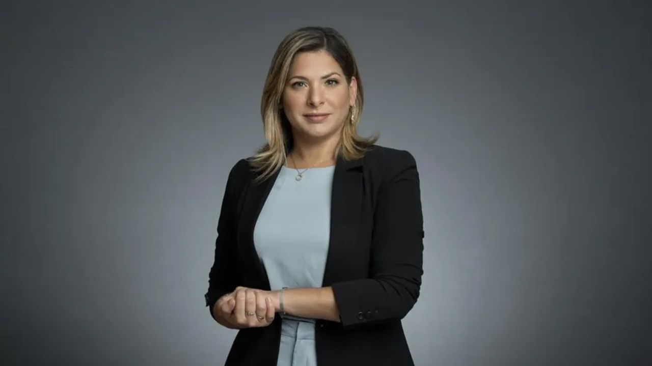Daniela Lima de roupa cinza e preta, posando com mãos unidas e sem sorrir