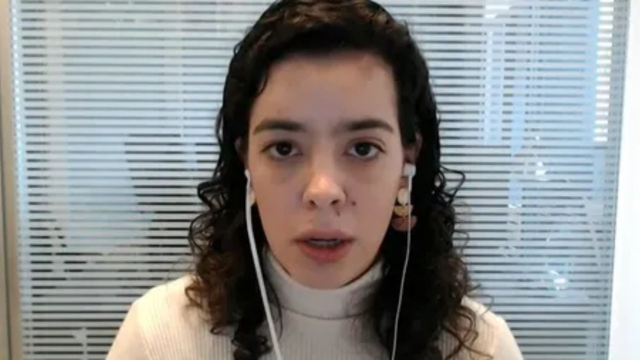 Maira Pinheiro de fones de ouvido e blusa clara, falando e olhando pra câmera