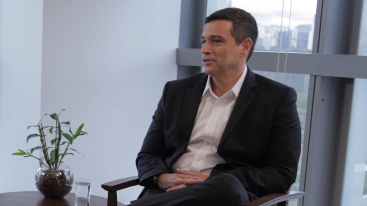 Roberto Campos Neto de roupa social, sentado com pernas cruzadas e falando, de perfil