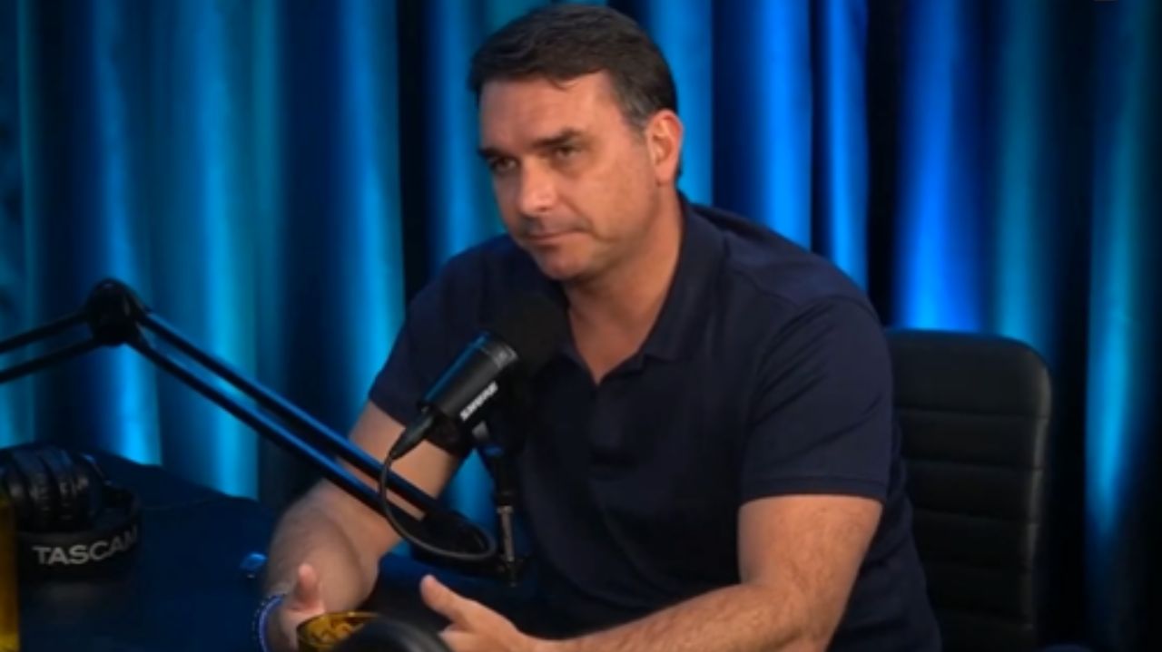 Flávio Bolsonaro falando em microfone, de camisa polo azul marinho