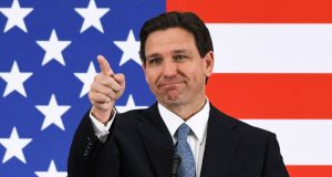 Ron DeSantis apontando pra frente com bandeira dos Estados Unidos ao fundo