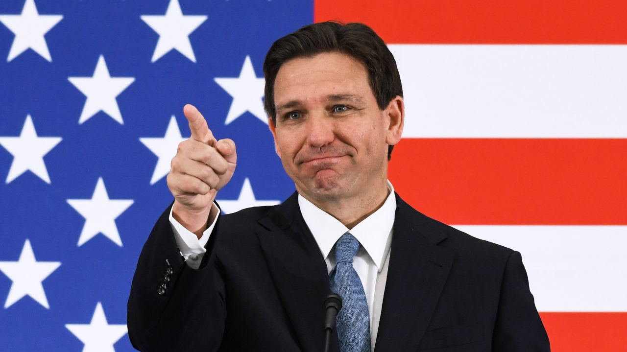 Ron DeSantis apontando pra frente com bandeira dos Estados Unidos ao fundo