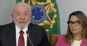 Lula falando em microfone ao lado de Janja, que está com roupa rosa e expressão séria