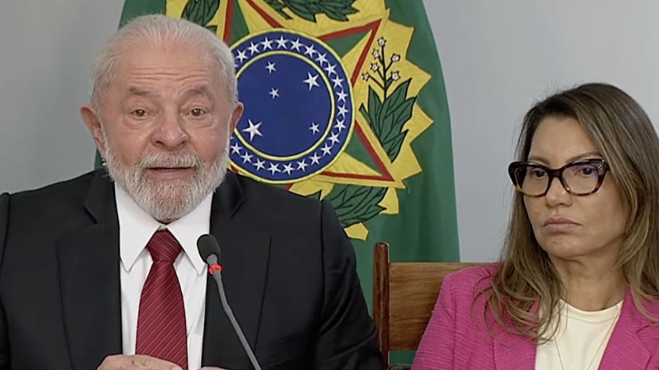 Lula falando em microfone ao lado de Janja, que está com roupa rosa e expressão séria