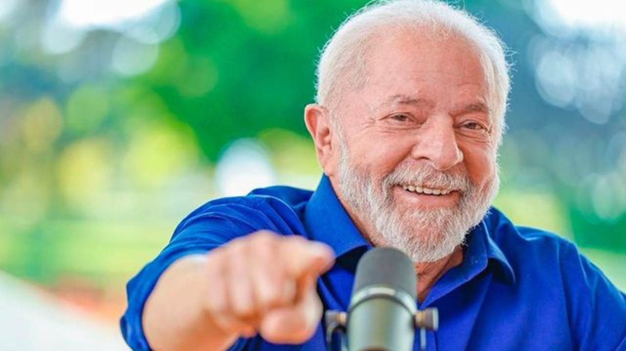 Lula de camisa social azul, sorrindo e apontando pra frente