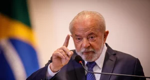 Lula falando em microfone e gesticulando, de terno e gravata, com bandeira do Brasil ao fundo