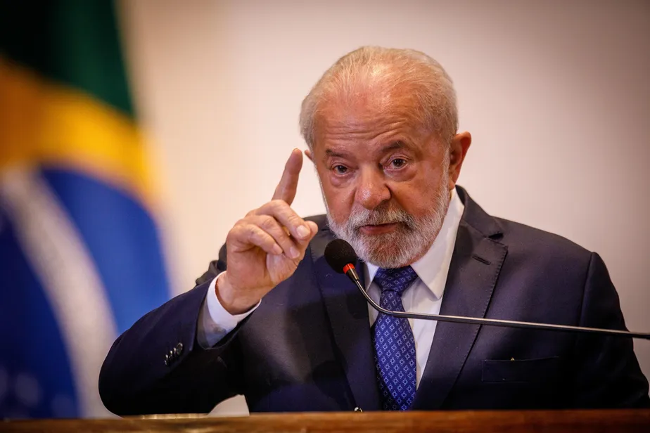 Lula falando em microfone e gesticulando, de terno e gravata, com bandeira do Brasil ao fundo