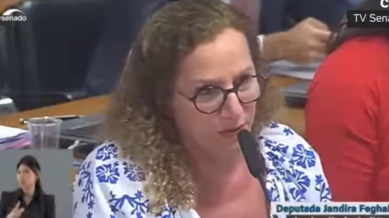 Jandira Feghali de óculos, com blusa branca e azul, falando em microfone