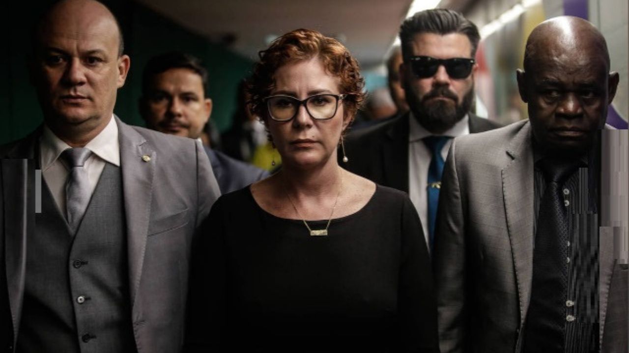 Carla Zambelli cercada, de óculos e roupa preta, com expressão séria