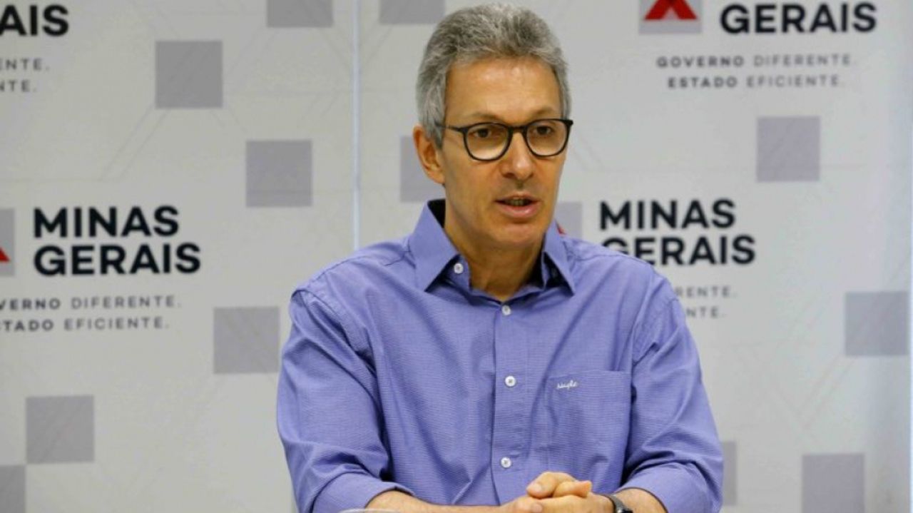 Romeu Zema de roupa social azul, falando, de óculos, com mãos unidas