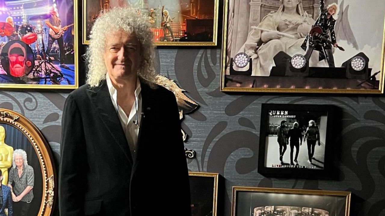 Brian May posando para foto perto de parede de quadros