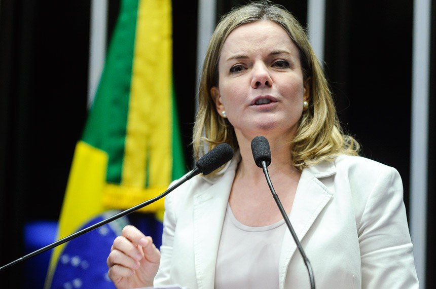 Gleisi Hoffmann falando em microfone e vestindo roupa clara, perto de bandeira do Brasil