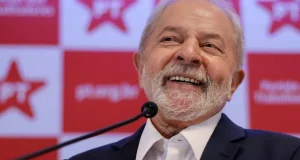Lula sorrindo com painel do PT ao fundo, perto de microfone