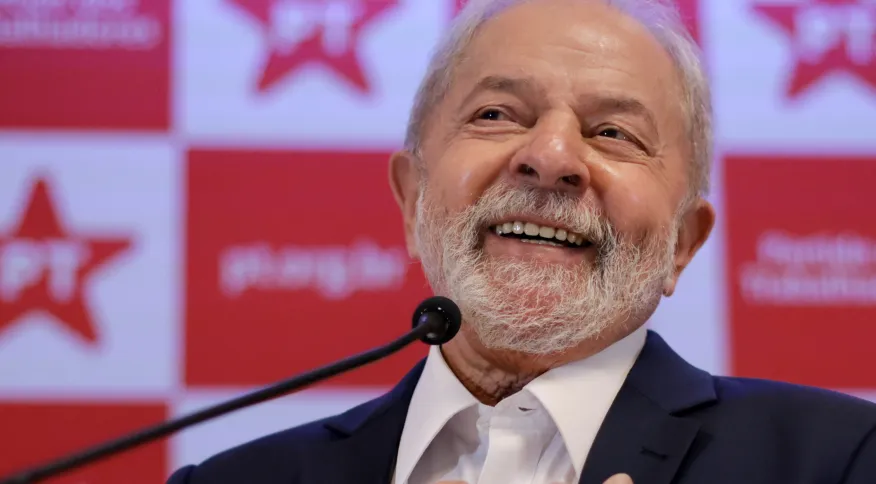 Lula sorrindo com painel do PT ao fundo, perto de microfone