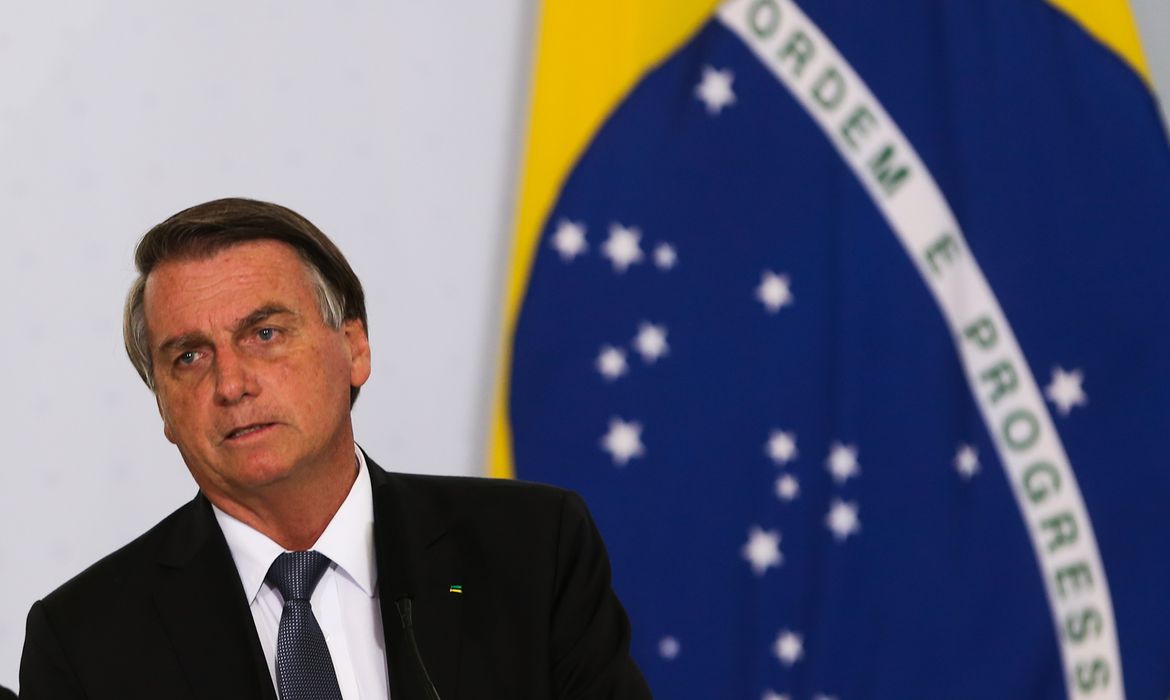 Jair Bolsonaro com bandeira do Brasil ao fundo e expressão de preocupação