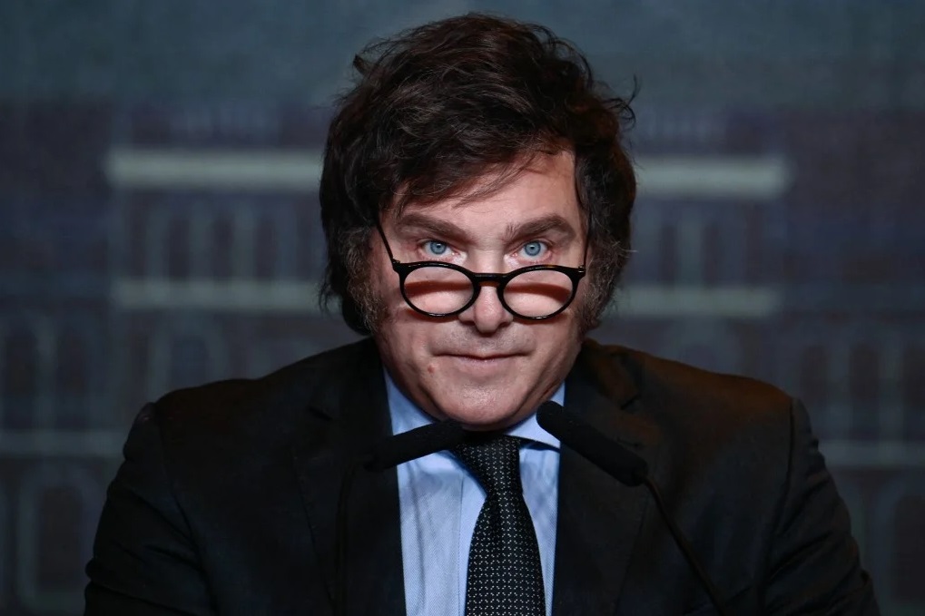 Javier Milei com expressão séria, de óculos, falando em microfone