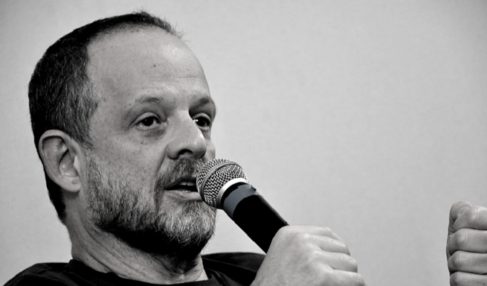 Breno Altman em foto em preto e branco, falando em microfone