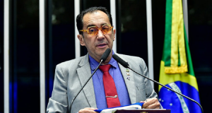 Jorge Kajuru com roupa e óculos coloridos, segurando camiseta e falando