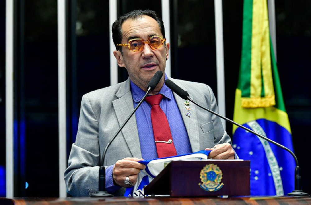 Jorge Kajuru com roupa e óculos coloridos, segurando camiseta e falando