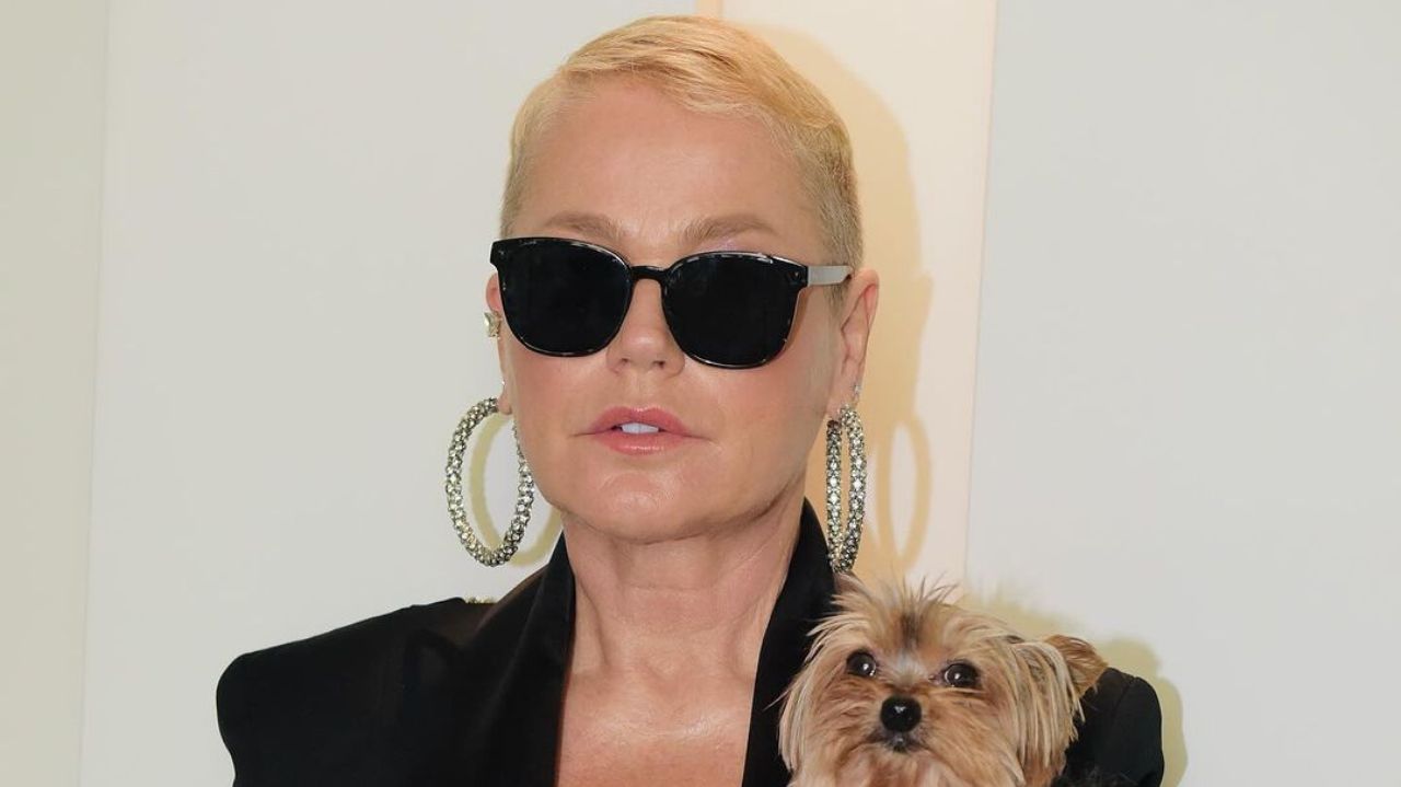 Xuxa de roupa preta, brincos de argola e óculos escuros, segurando cachorrinha
