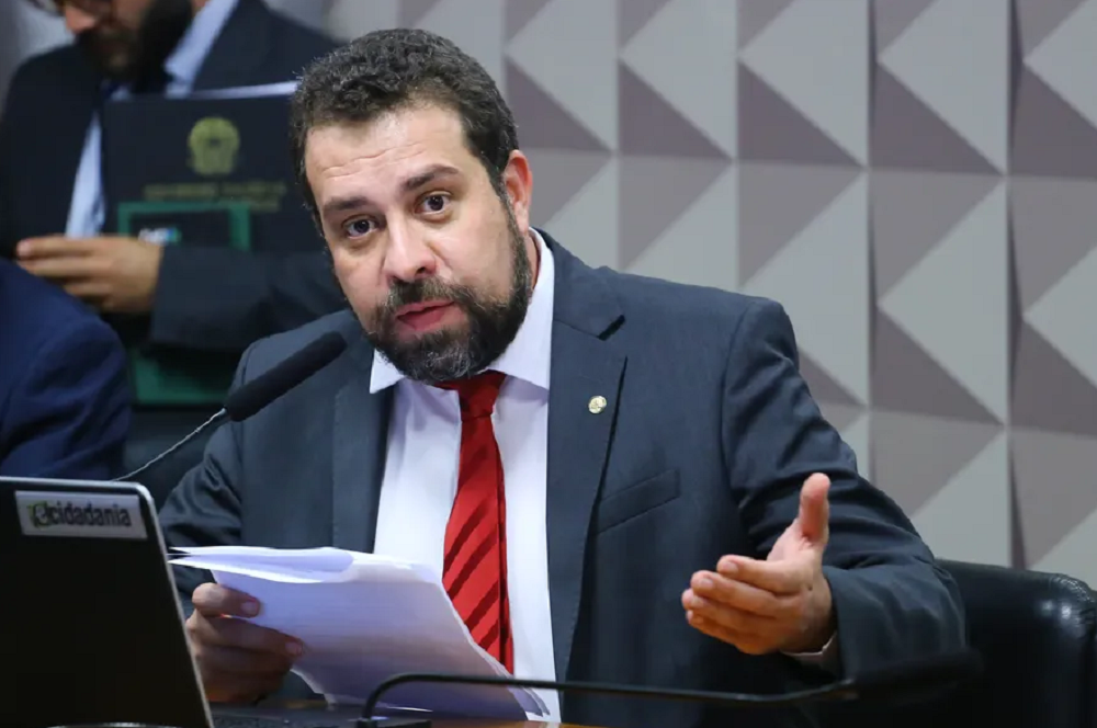 Guilherme Boulos falando em microfone, segurando papel e gesticulando