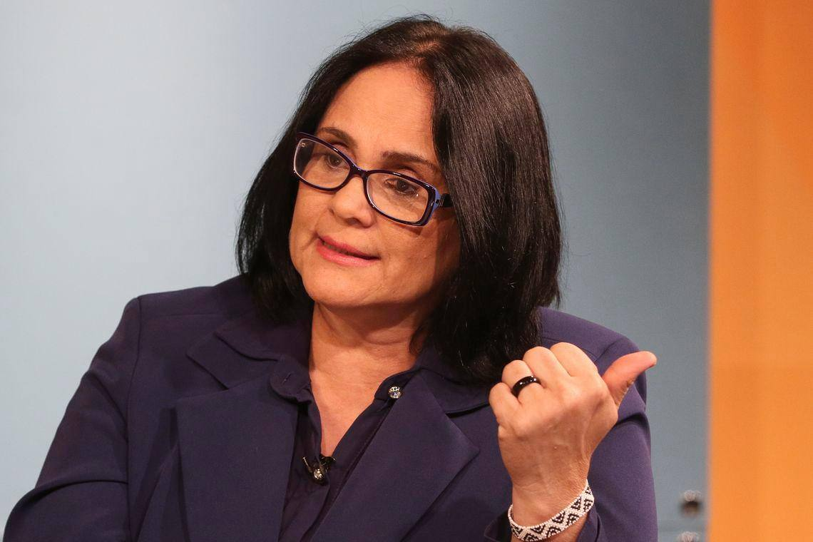 Damares falando, vestindo roupa social azul escura e apontando pra trás, de óculos