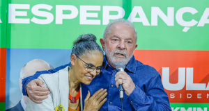 Marina Silva e Lula abraçados, ela sorrindo e ele falando em microfone