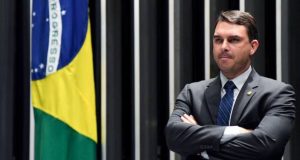 Flávio Bolsonaro com expressão de satisfação perto de bandeira do Brasil