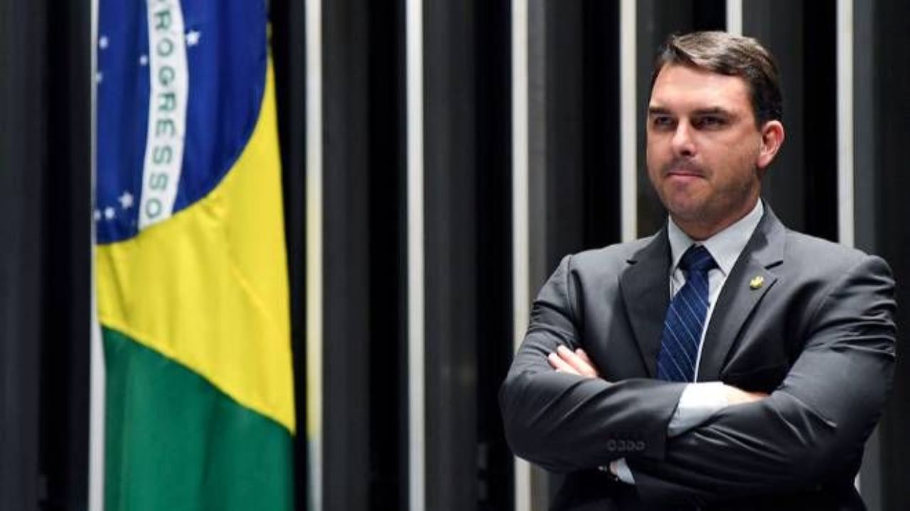 Flávio Bolsonaro com expressão de satisfação perto de bandeira do Brasil