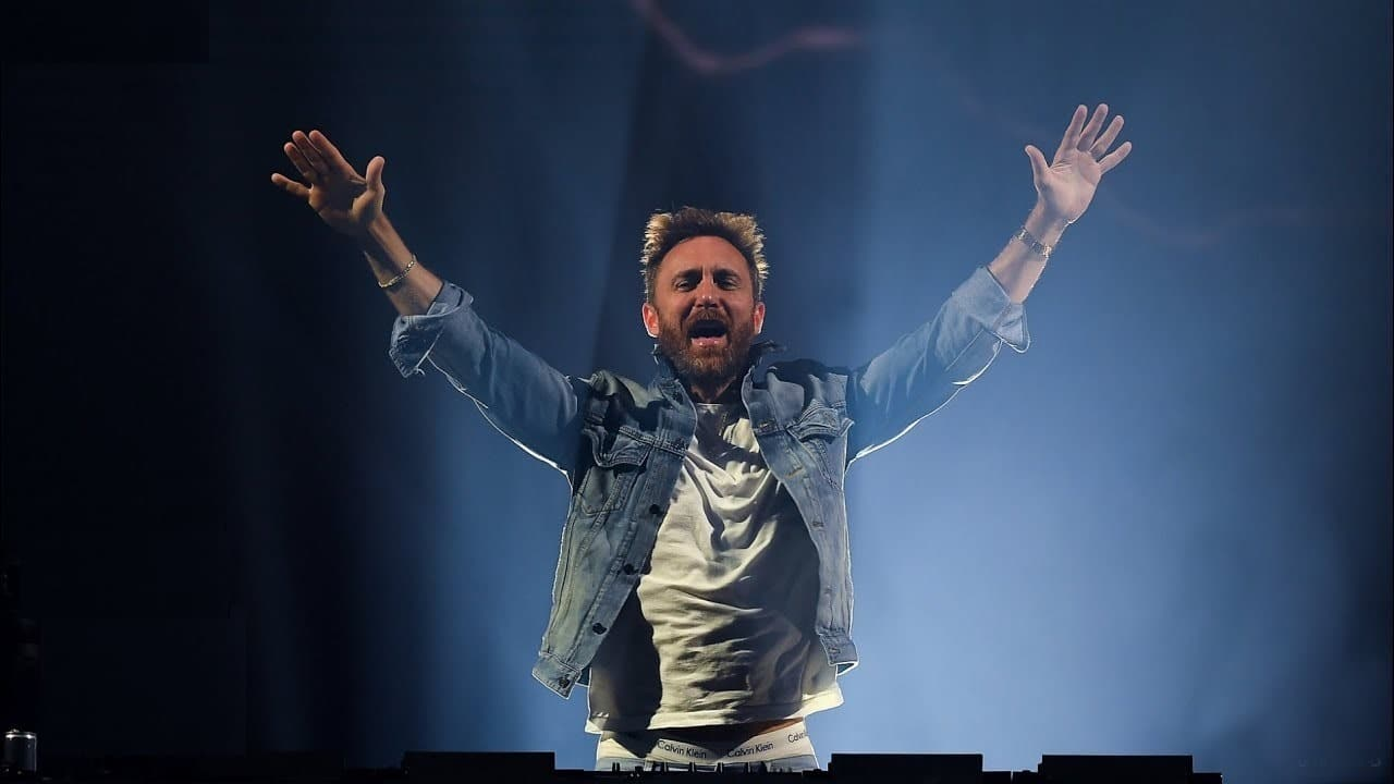 David Guetta em show, com jaqueta jeans e os braços abertos