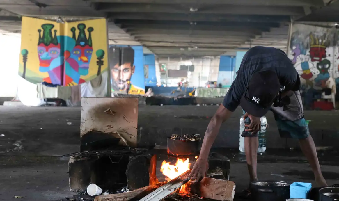 Homem abaixado, de roupa e boné pretos, cozinhando embaixo de viaduto