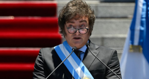 Javier Milei falando em microfone, com faixa presidencial e óculos