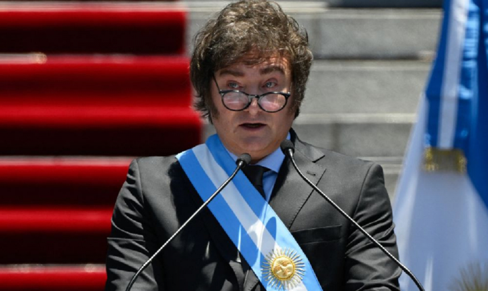 Javier Milei falando em microfone, com faixa presidencial e óculos