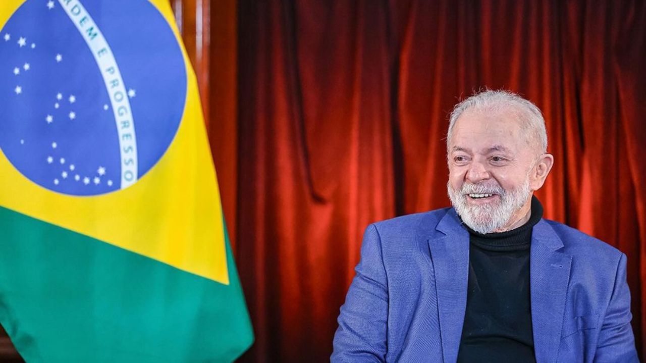 Lula sorrindo e olhando para o lado, com bandeira do Brasil ao fundo