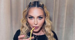 Madonna segurando copo e bebendo em canudo, de roupa preta e cabelo solto