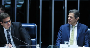 Fernando Haddad olhando para Roberto Campos Neto, que está falando em microfone