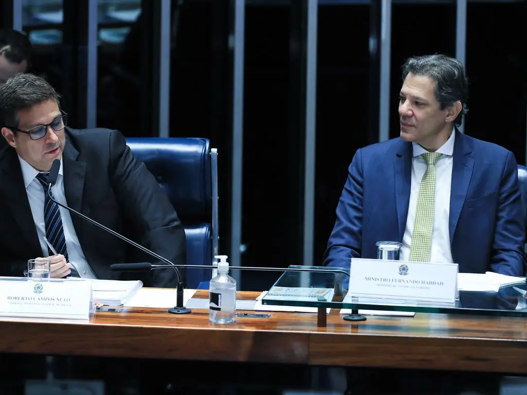 Fernando Haddad olhando para Roberto Campos Neto, que está falando em microfone