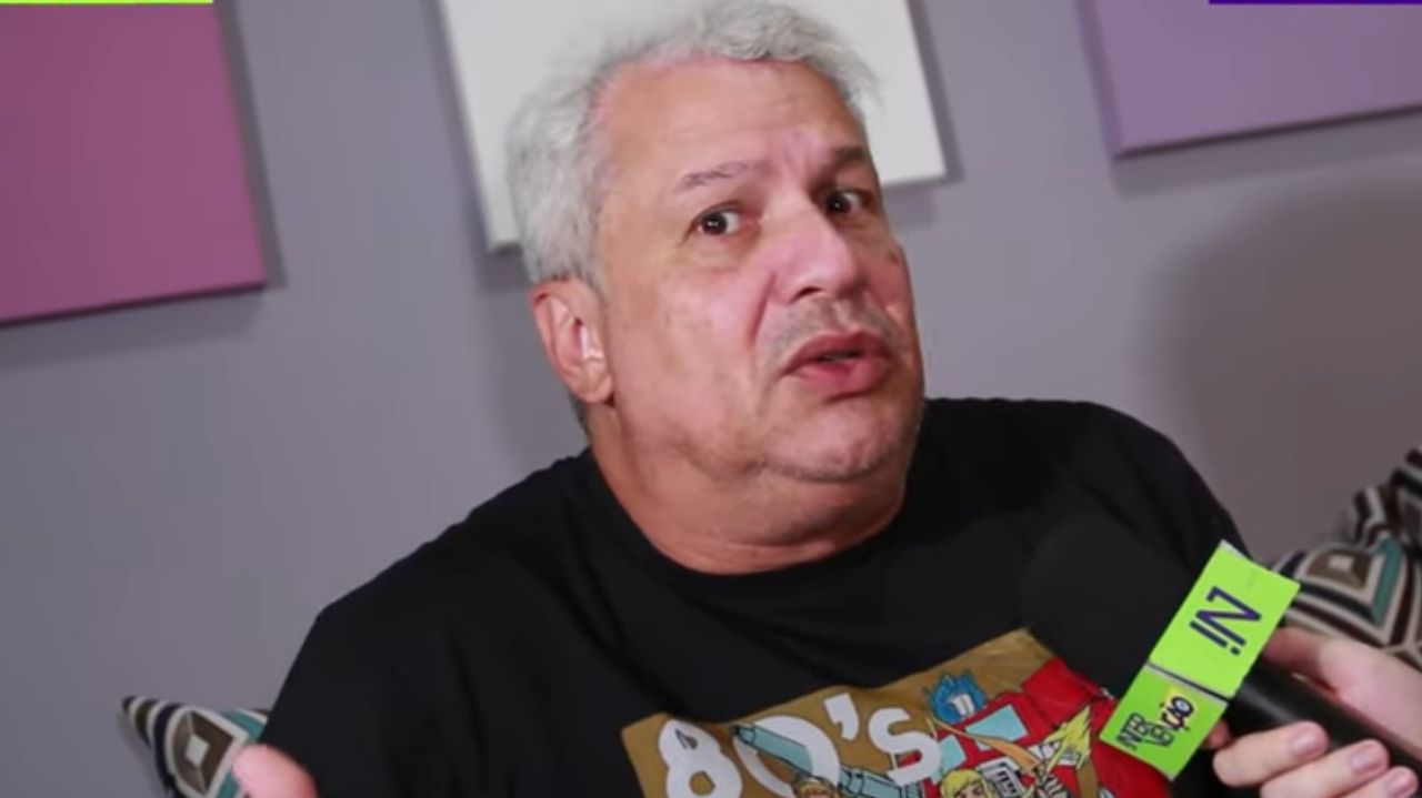 Sikêra Jr. de camiseta preta, falando em microfone, com expressão confusa