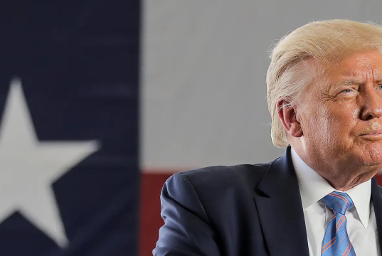 Donald Trump olhando para o lado perto de bandeira dos EUA