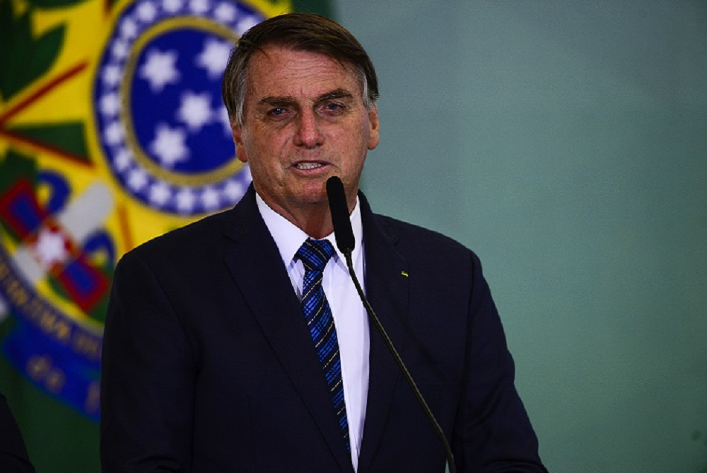 Jair Bolsonaro de terno e gravata, falando em microfone, com bandeira atrás