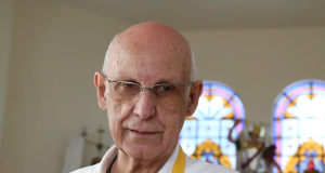 Padre Júlio Lancellotti com roupa branca e avental amarelo, com expressão séria e óculos