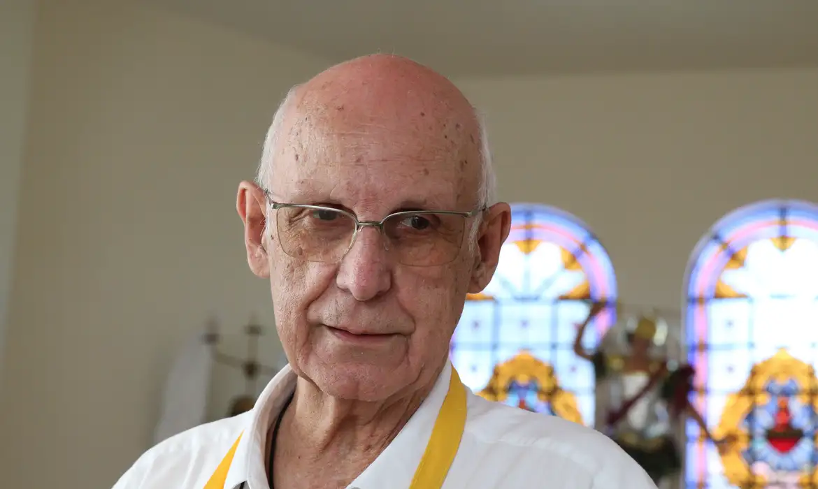 Padre Júlio Lancellotti com roupa branca e avental amarelo, com expressão séria e óculos