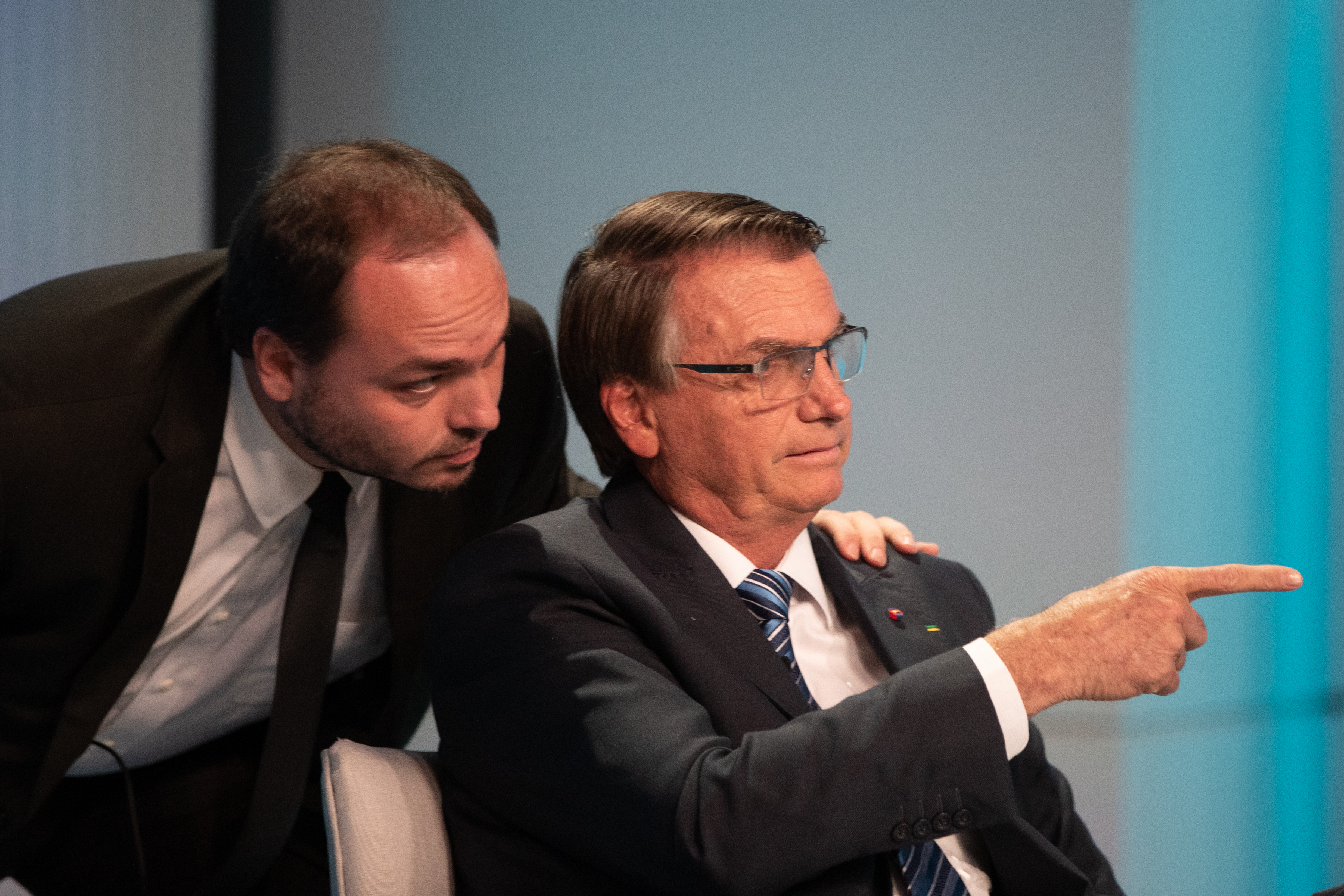 Carlos Bolsonaro falando com Jair Bolsonaro, agachado