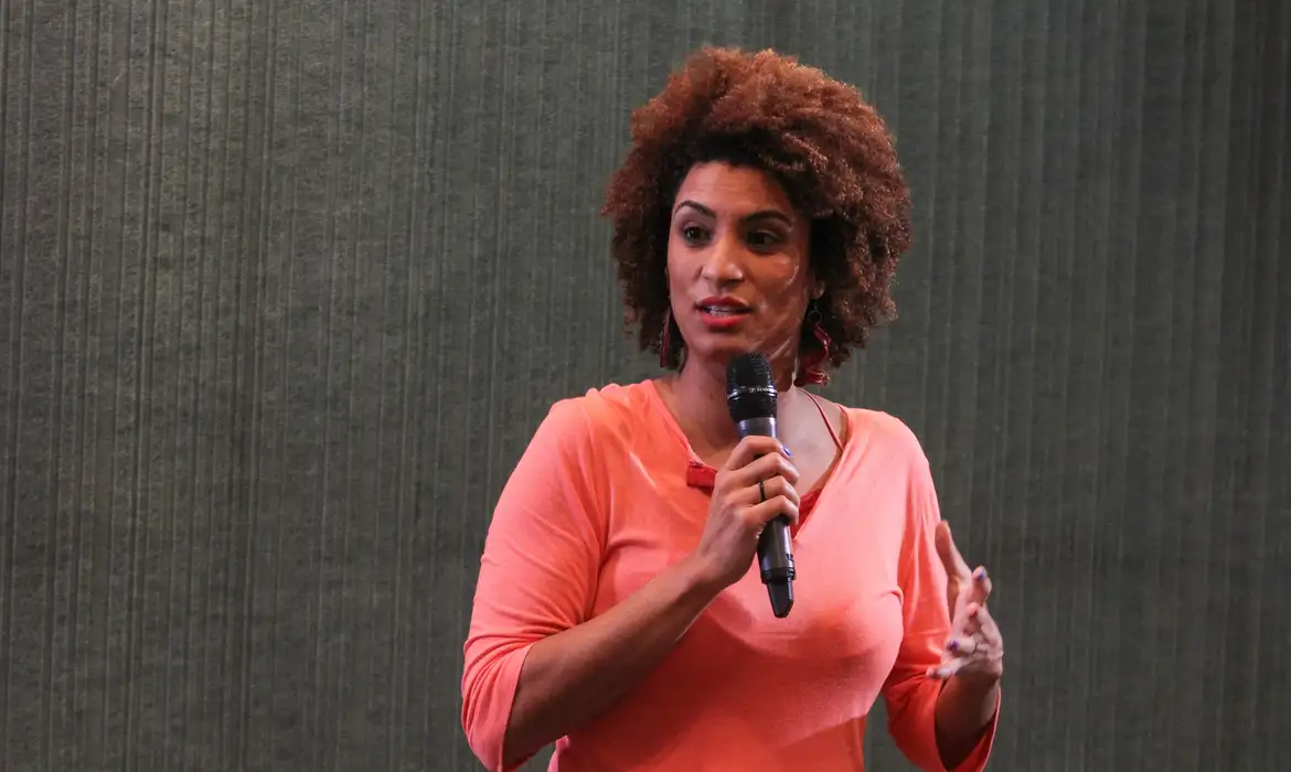 Marielle Franco de roupa vermelha, falando em microfone e olhando para o lado