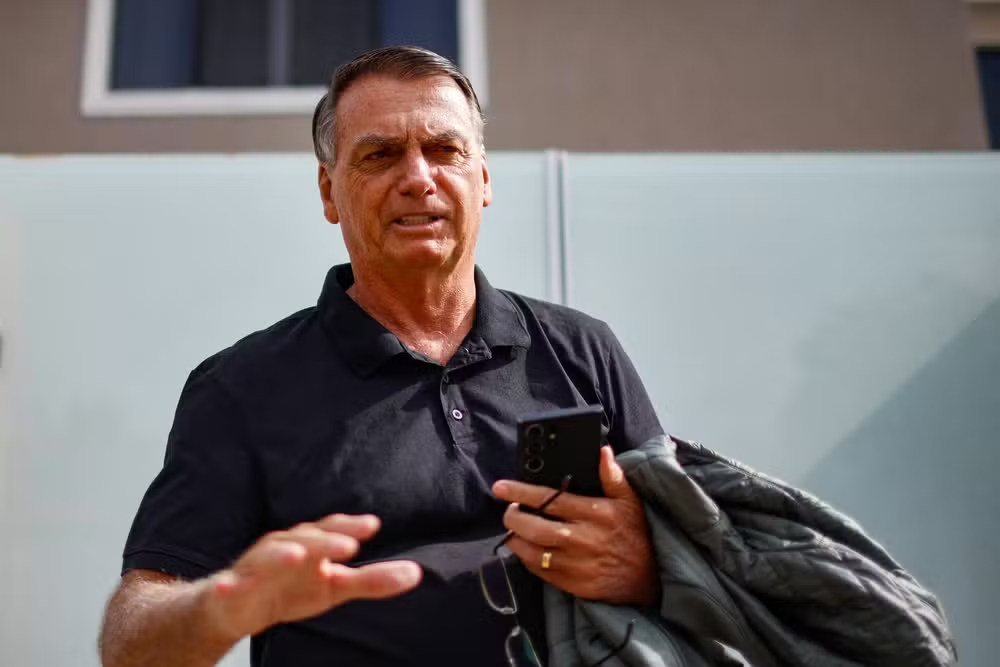 Jair Bolsonaro com camisa polo escura, falando e pedindo calma, carregando celular, óculos e casaco
