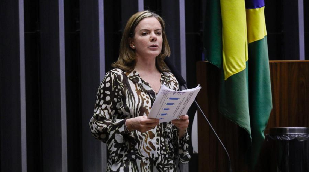 Gleisi Hoffmann falando em microfone com expressão séria, segurando papel, com roupa estampada